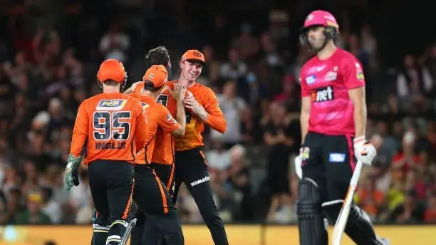 Cricketers in orange celebrate while a pink-clad batter stands nearby during a match.