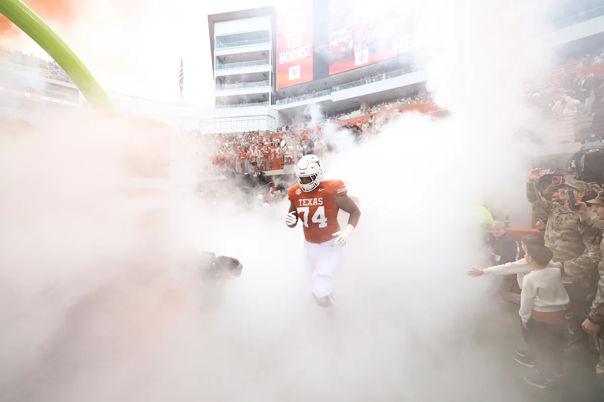 Football player in Texas uniform emerges through smoke as fans line the stadium tunnel.