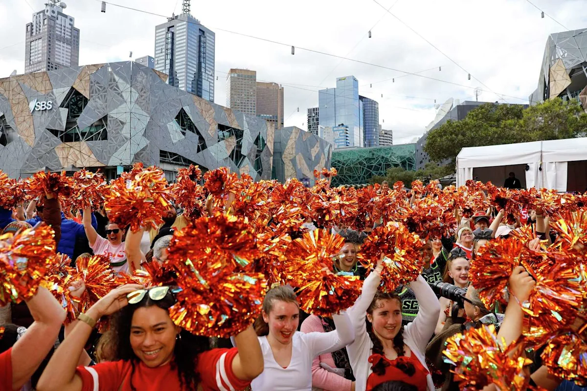 Crowd waves orange pom-poms outside a geometric stadium surrounded by Melbourne skyscrapers.