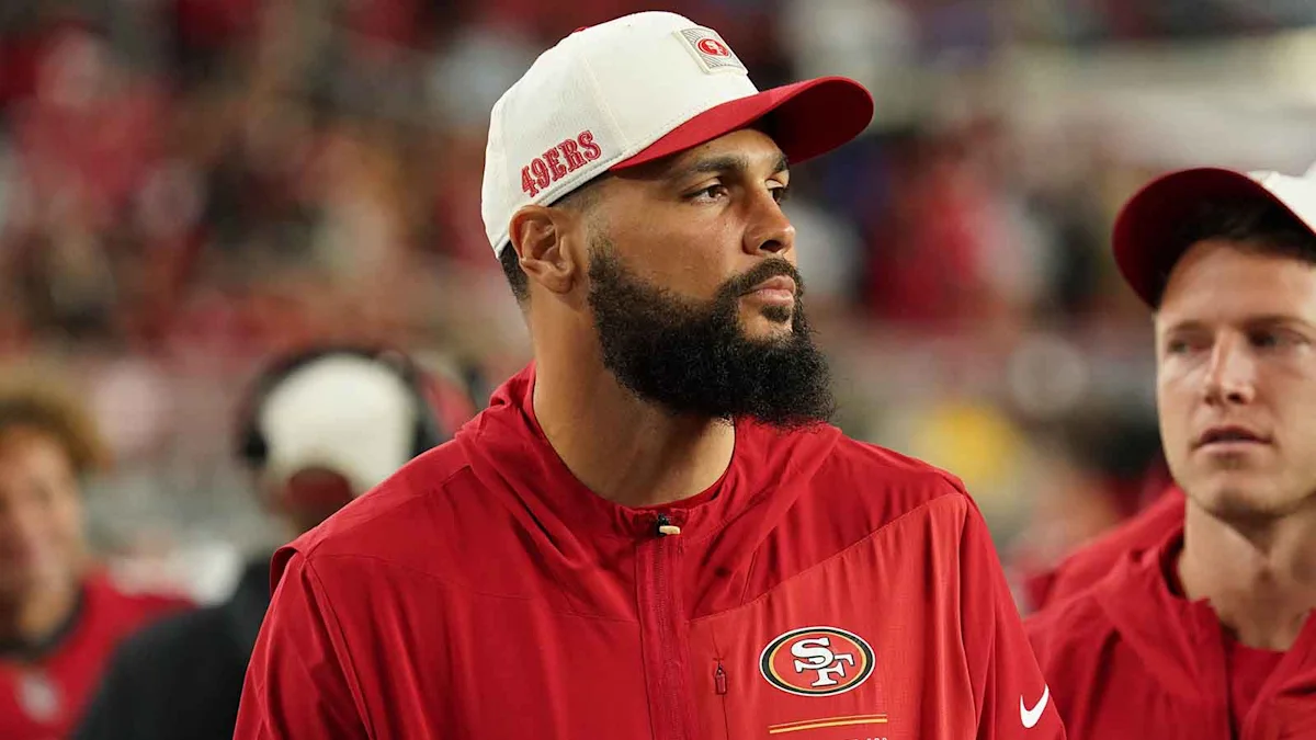 Bearded football player in red 49ers attire and white cap standing on sideline