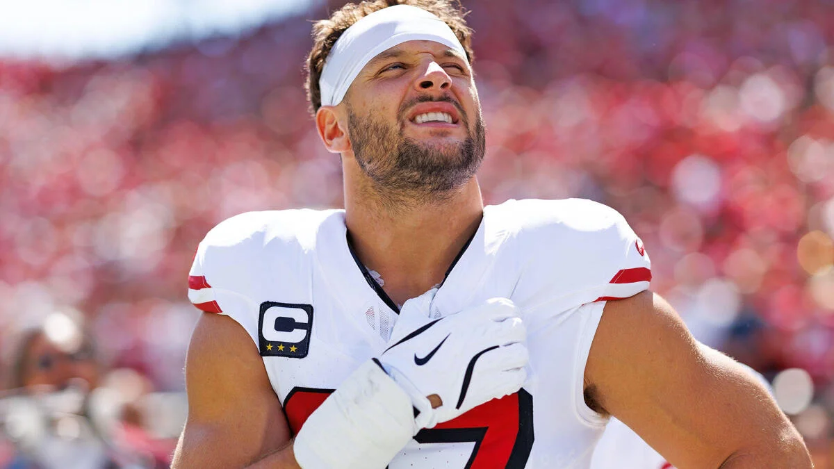 Football player in a white uniform and cap raises a gloved hand against a blurred stadium crowd.