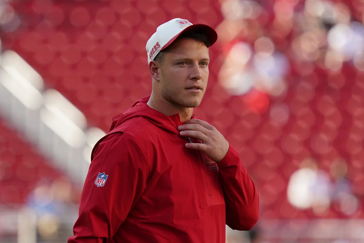 Football player in a red jacket and white 49ers cap stands before red stadium seats