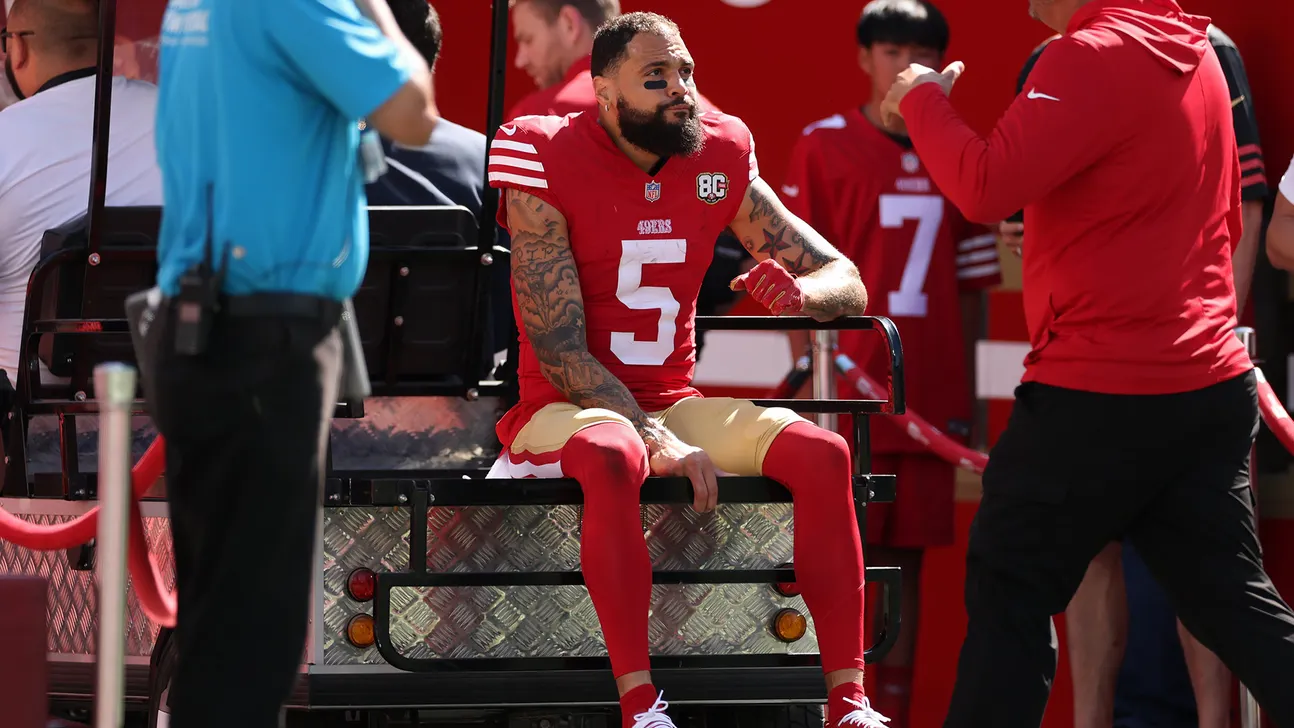 A football player in a red number 5 jersey sits on a cart as staff stand nearby.