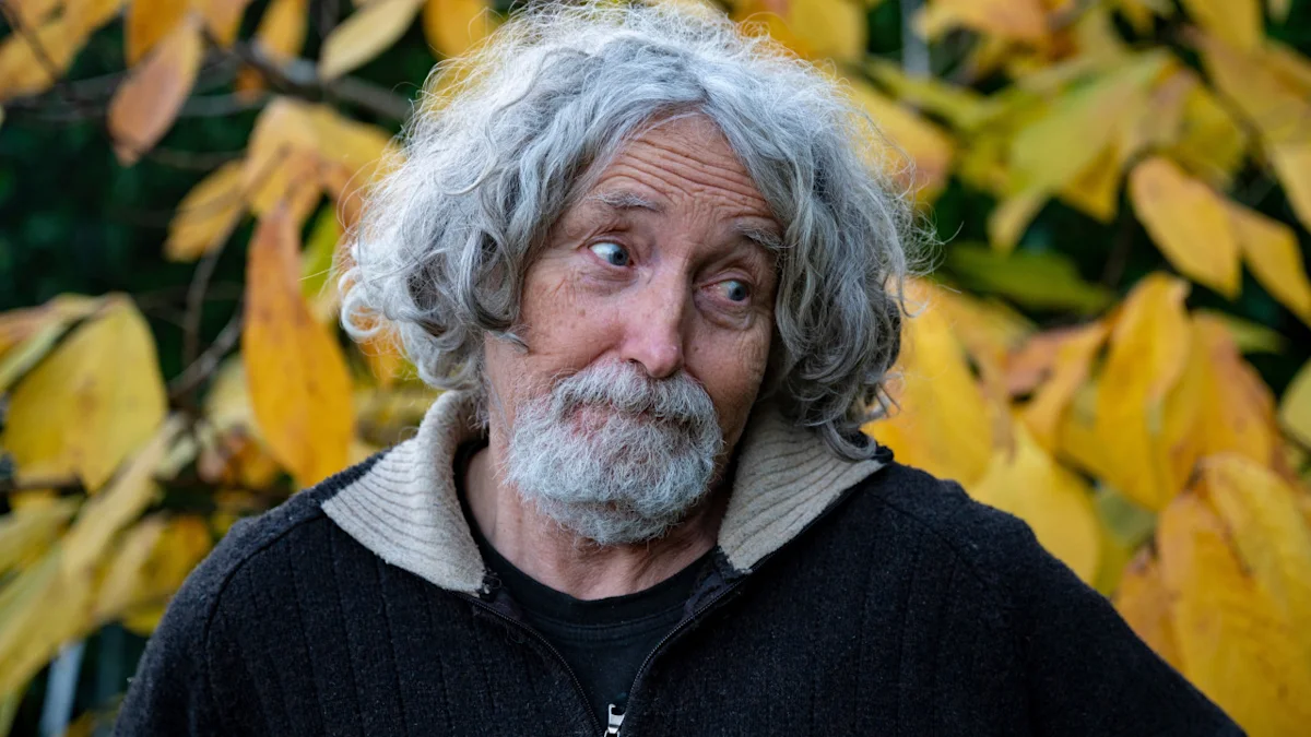 Gray-haired man with a mustache wearing a dark sweater, posing outdoors among yellow autumn leaves.