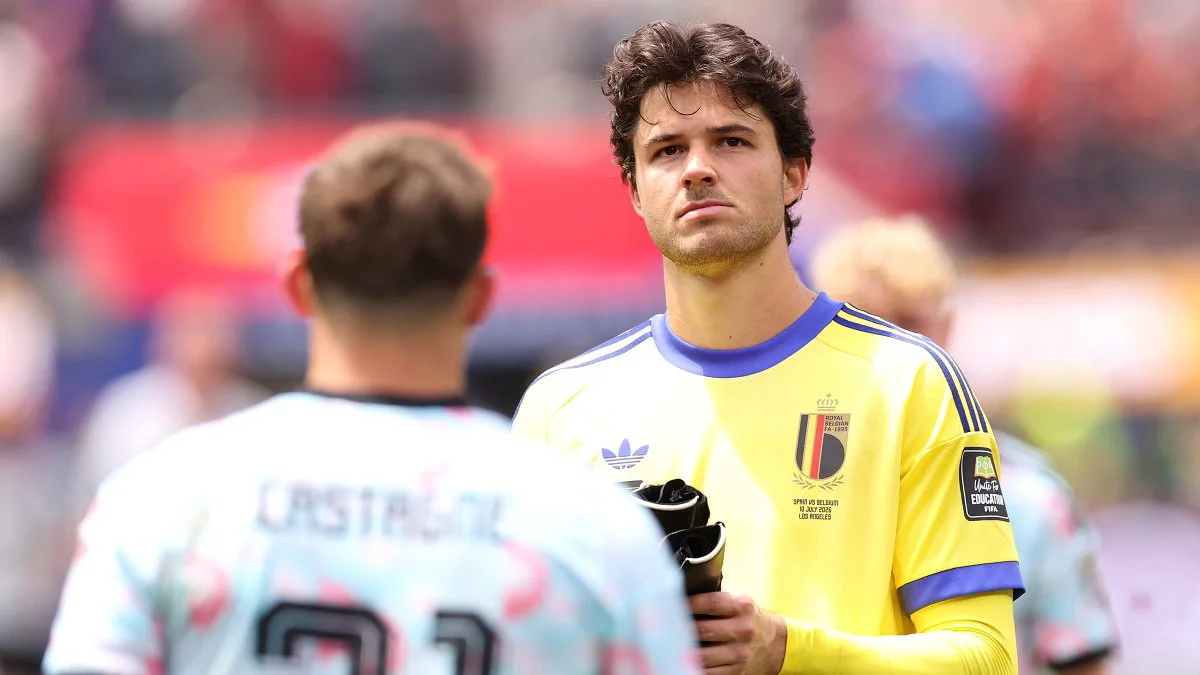 A goalkeeper in a yellow Belgium jersey holds gloves while facing an opponent in a pale blue kit.