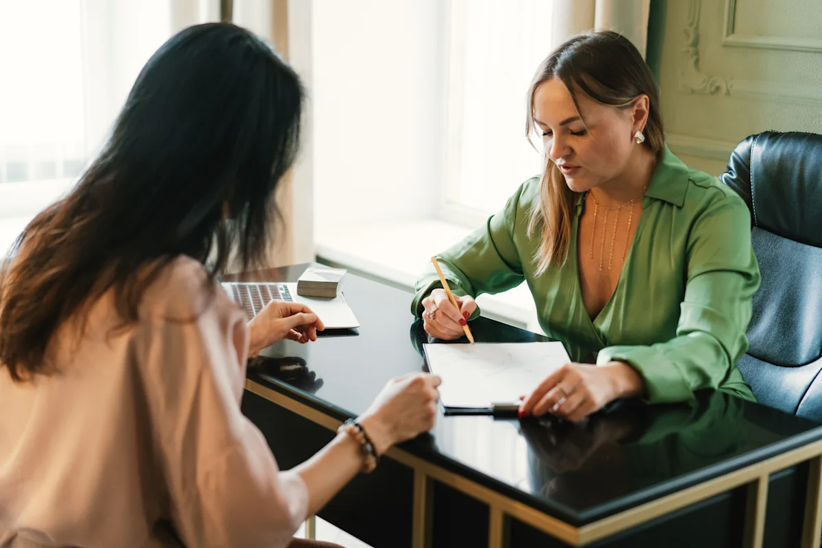 Two women discuss paperwork at a glass desk, with one writing in a notebook.