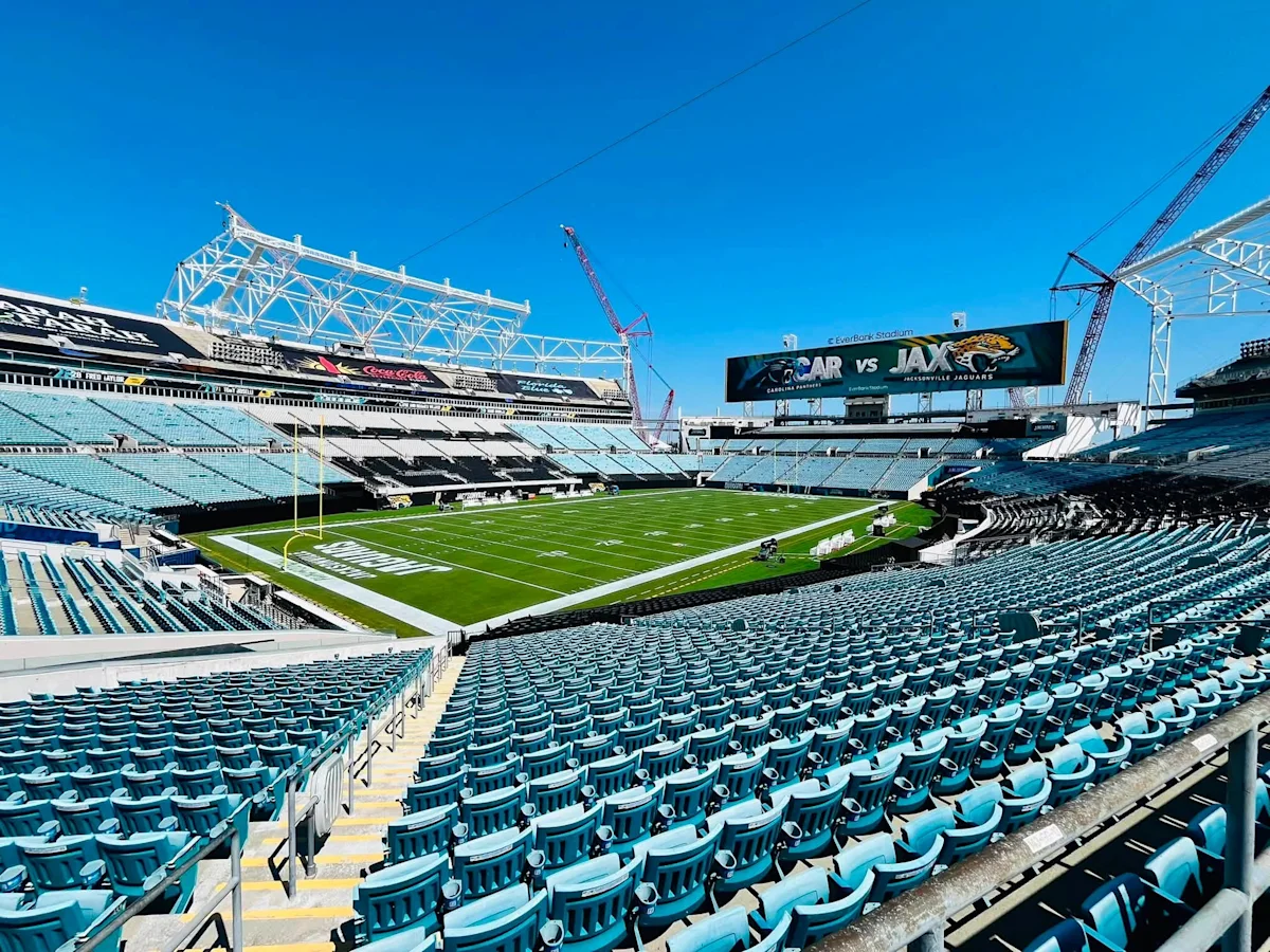 Empty football stadium with turquoise seats, green field, cranes, and a scoreboard showing CAR versus JAX.
