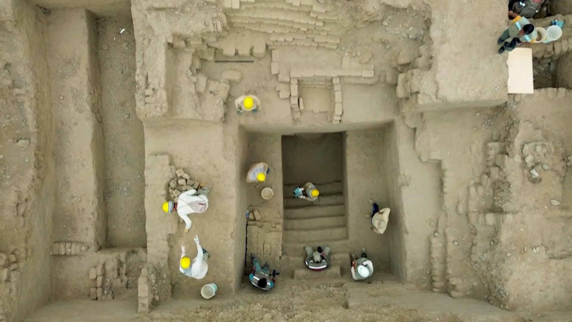 Aerial view of workers excavating a stepped earthen archaeological structure with exposed walls and chambers.