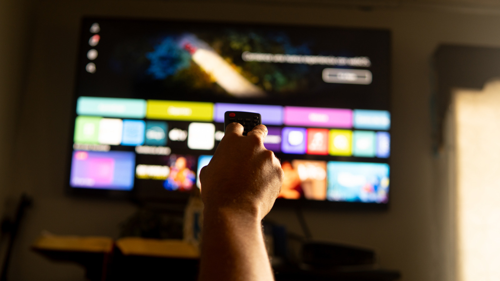 Hand holding a remote control toward a blurred television displaying colorful streaming service tiles.