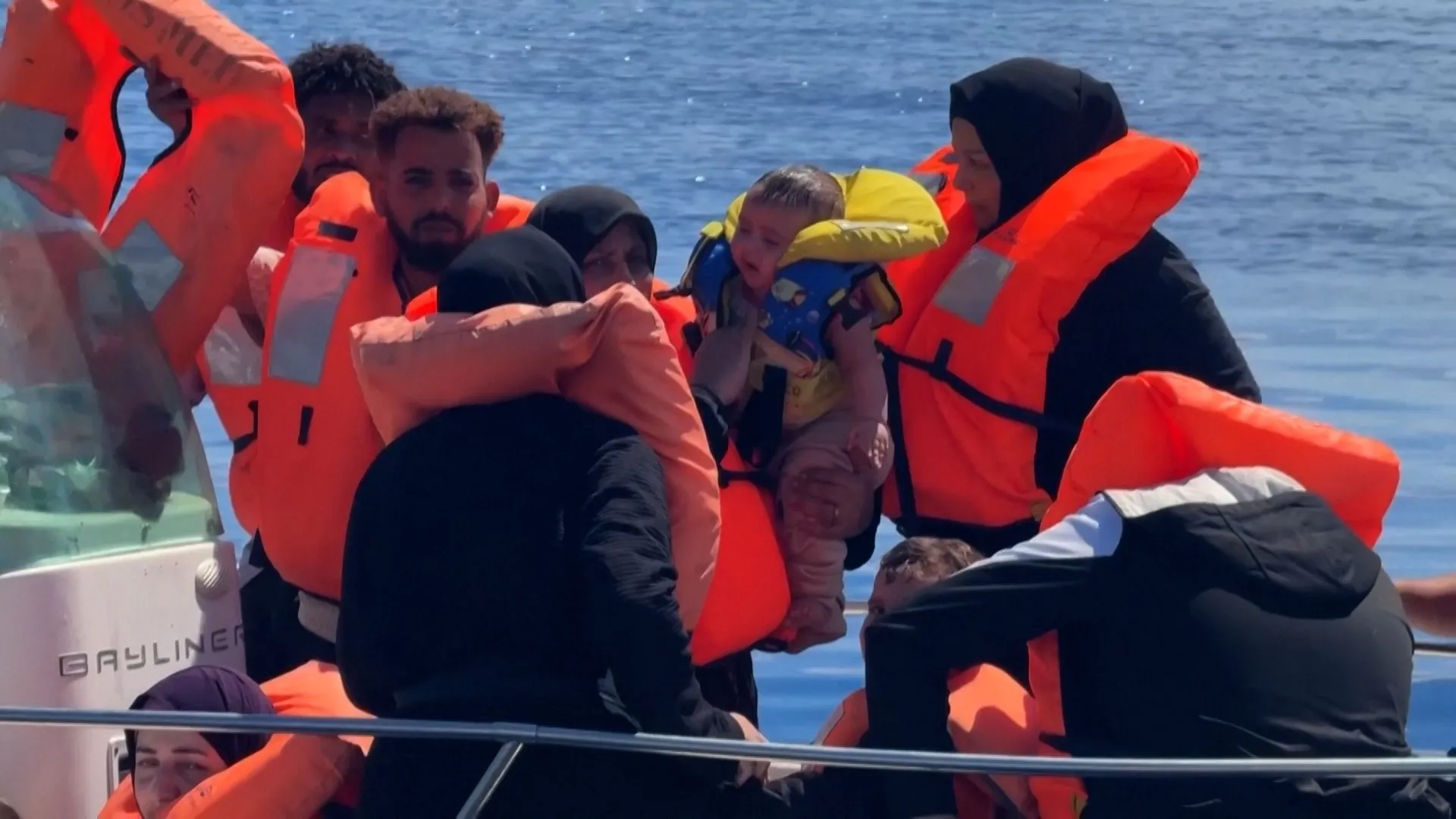Rescue workers in orange life jackets help adults and children aboard a boat at sea.