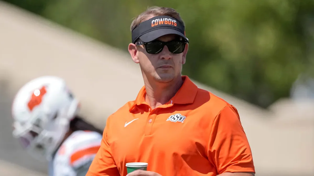 Man in orange Oklahoma State polo and visor stands holding a cup near football players
