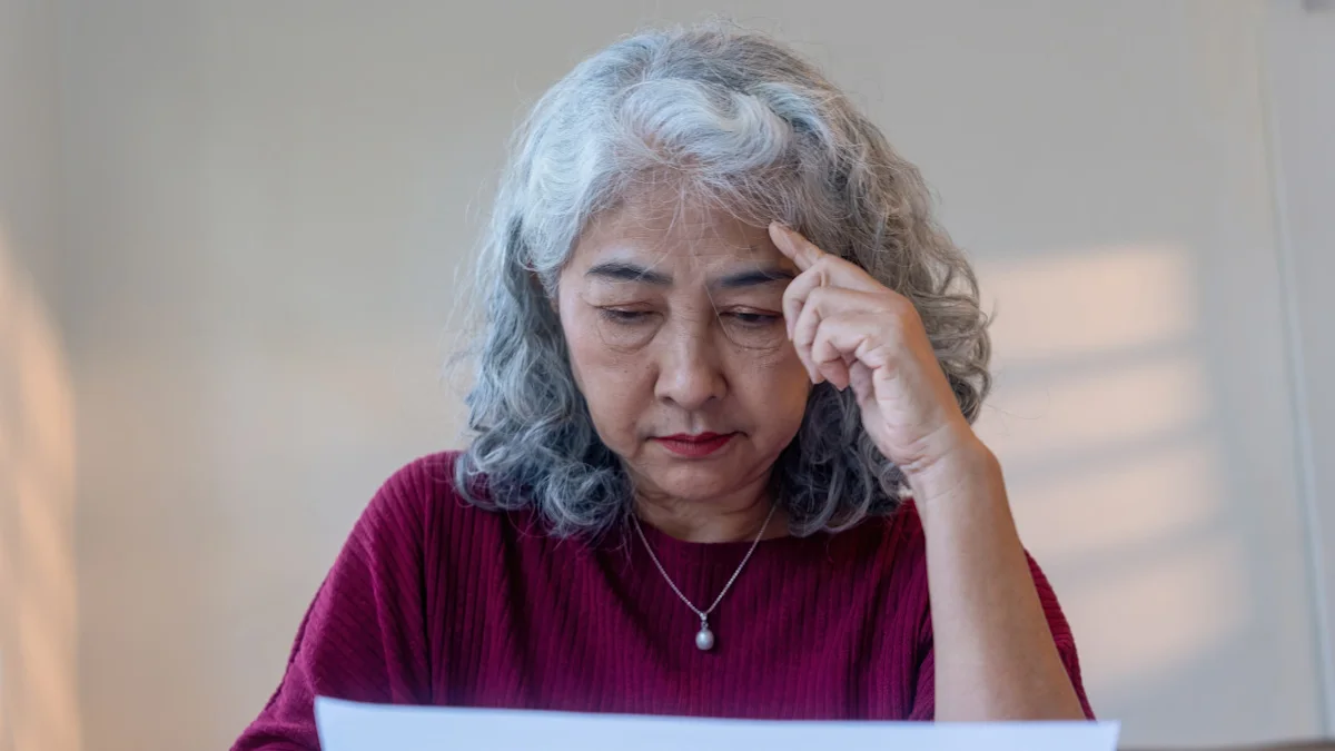 Gray-haired woman in burgundy sweater looks down at a document, touching her forehead.