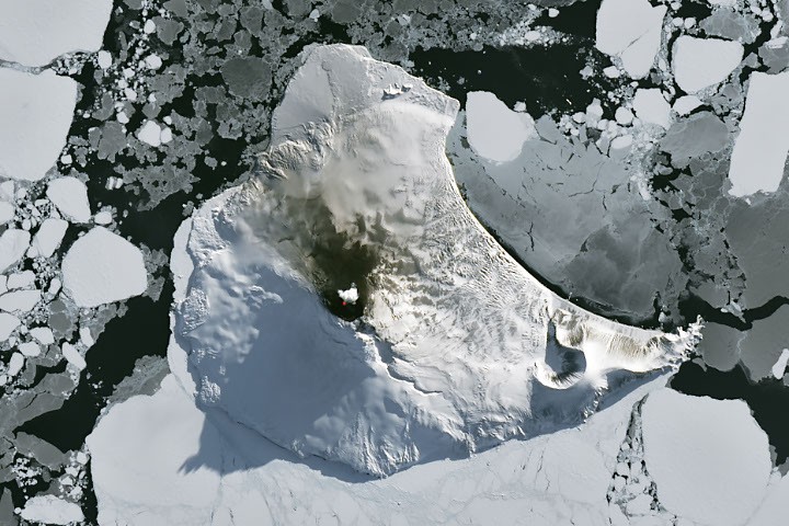 A snow-covered mountainous landform surrounded by dark water and fragmented sea ice