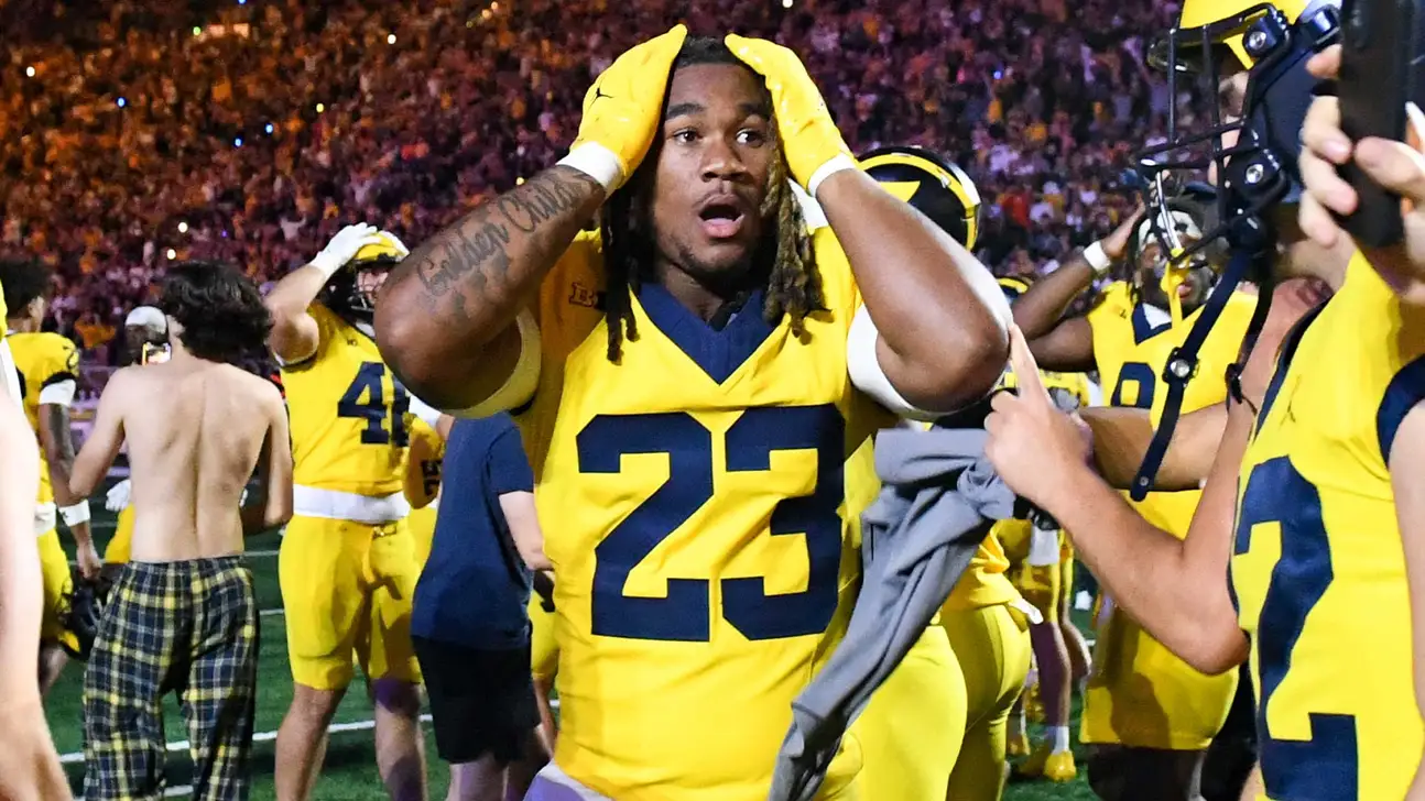 Football player in maize and blue holds his head amid teammates and a crowded field.