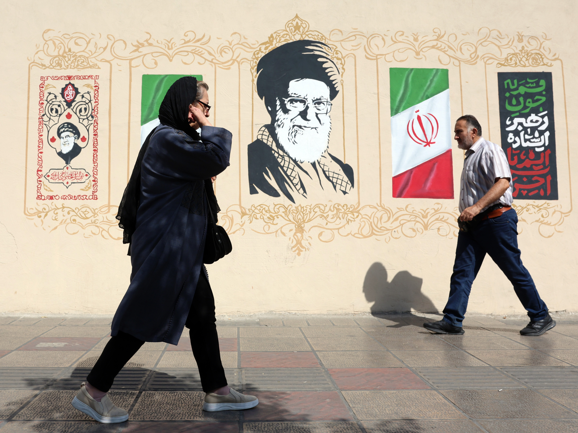 Two pedestrians walk past a mural depicting Iranian leaders, national symbols and Persian calligraphy.