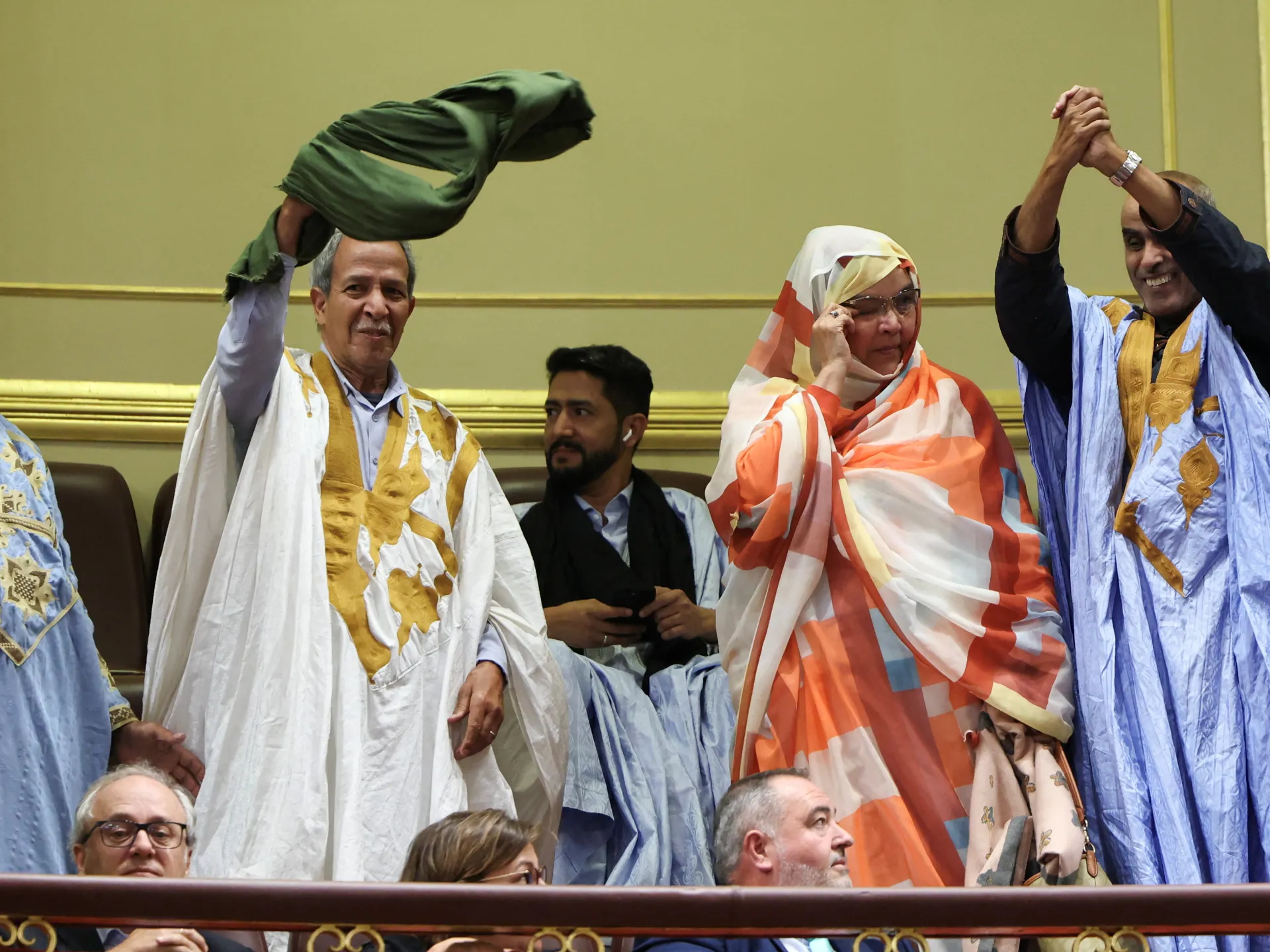 People in colorful traditional robes stand and raise their hands inside an assembly chamber.