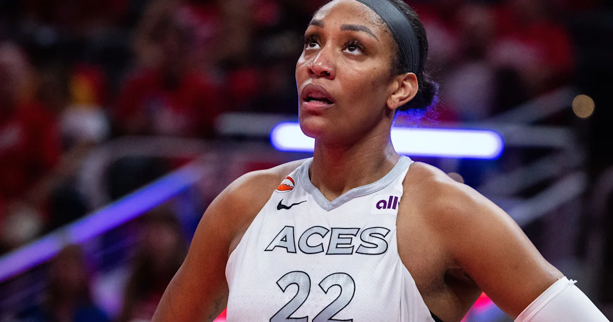 Basketball player in a white Aces jersey looks upward during a game, with a blurred arena crowd behind her.