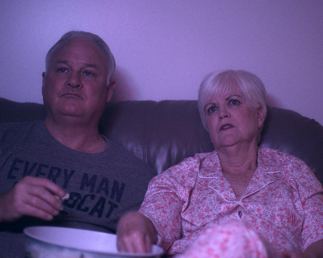 Two adults sit on a sofa in dim purple light, with one holding a bowl.