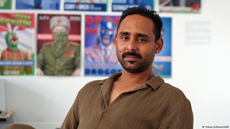 Man with mustache seated indoors before colorful posters, wearing a brown shirt and looking toward camera