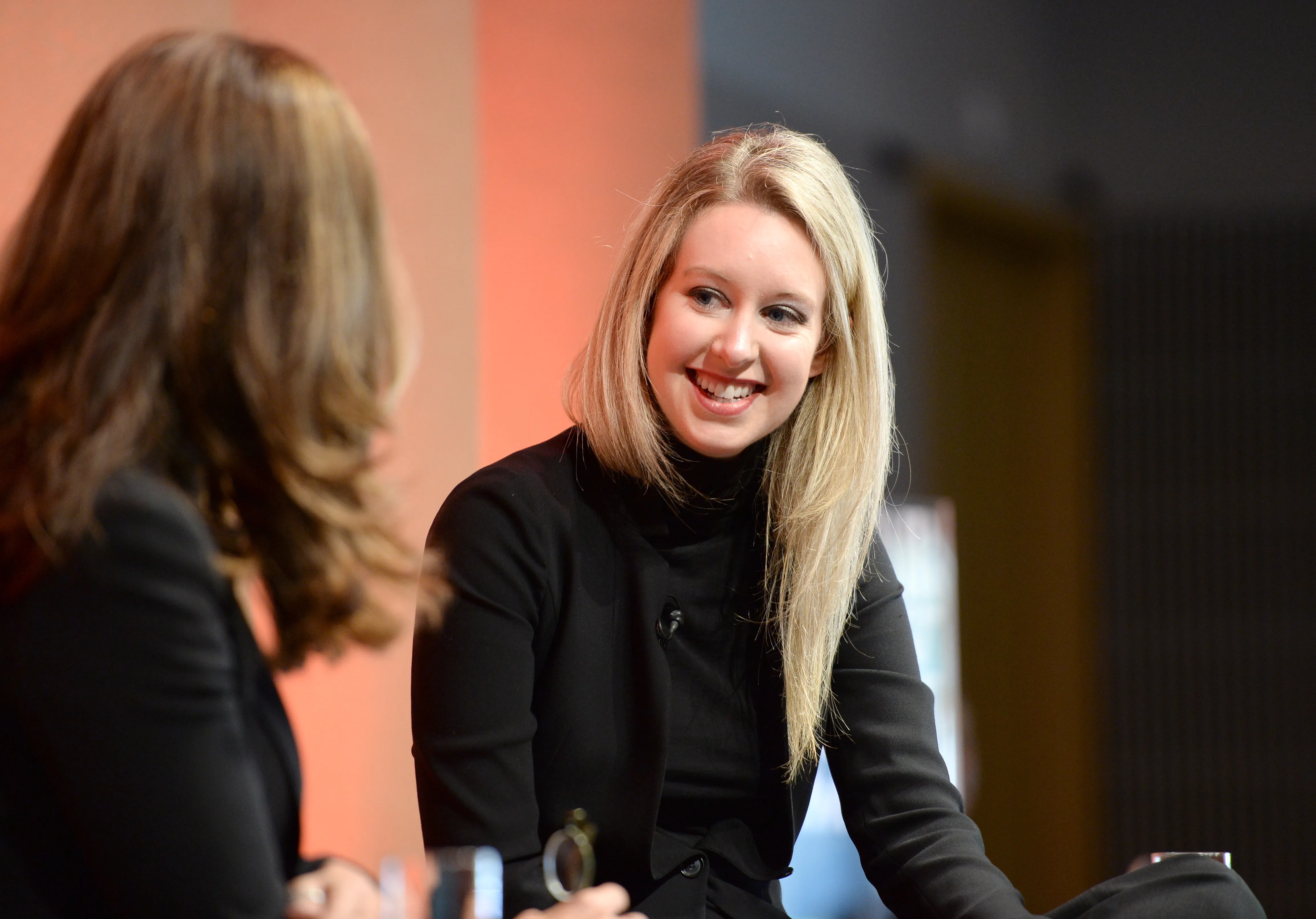 Smiling blonde woman in black speaks with another woman in a warmly lit interior.