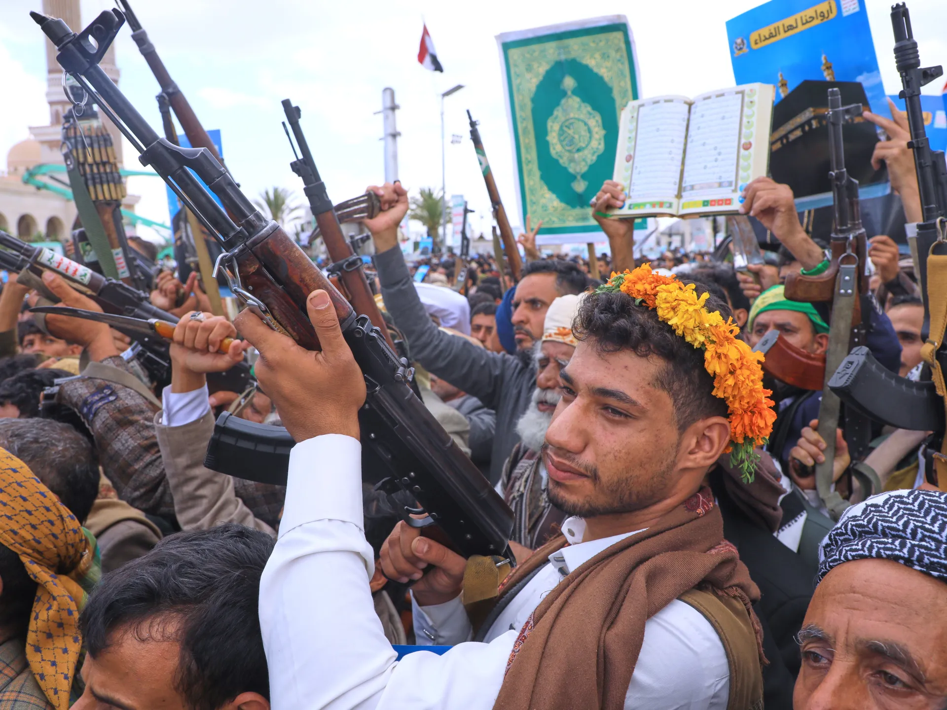 Crowd of armed men raising rifles, with an open Quran and mosque visible behind them