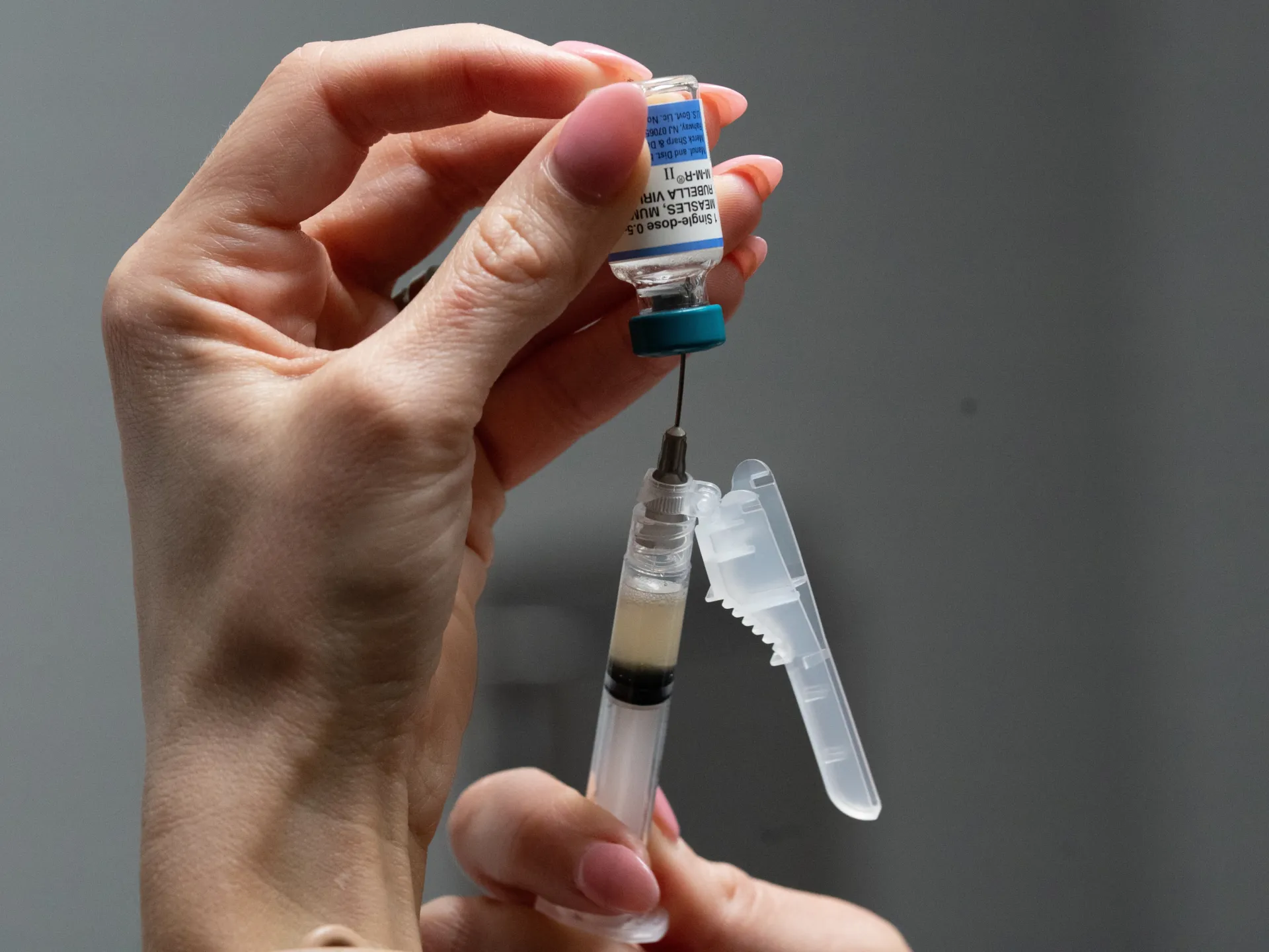 Hand holding a vaccine vial above a syringe and needle against a gray background