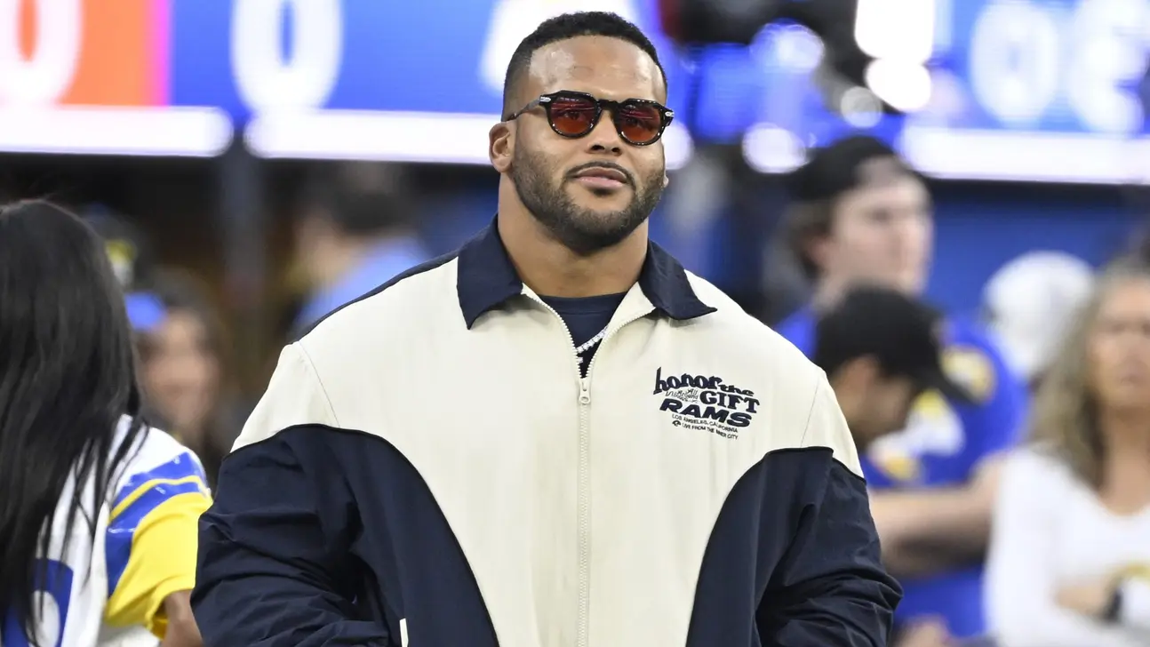 Man wearing sunglasses and a cream-and-navy jacket stands amid a football crowd.