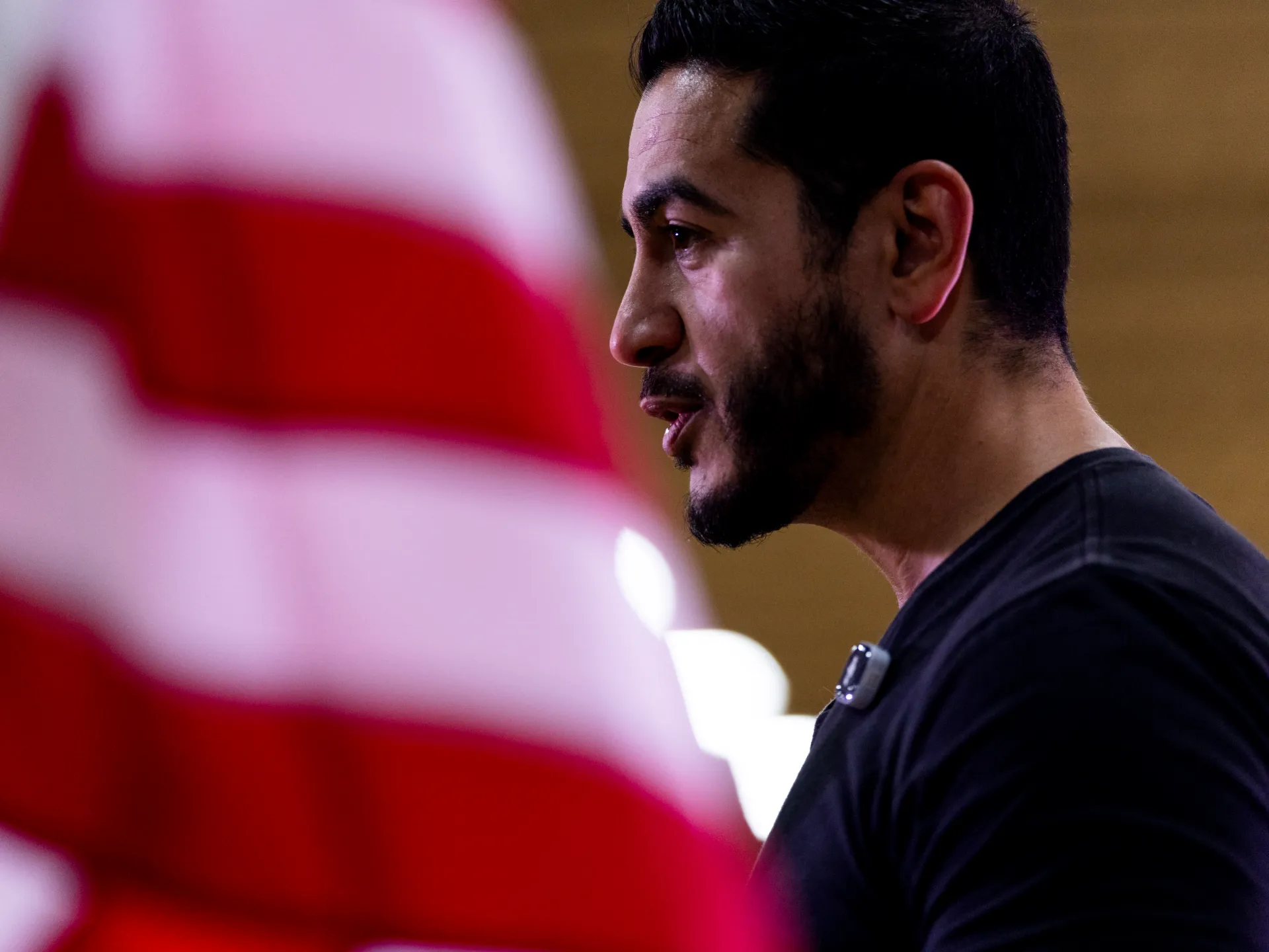Bearded man in profile wearing black shirt, with a blurred red-and-white foreground