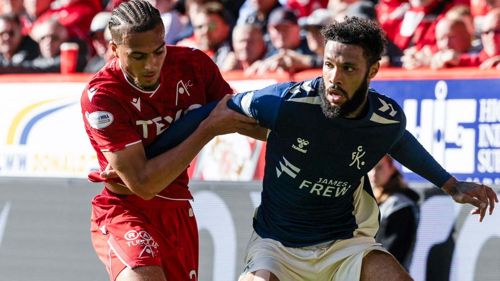 Two footballers compete for possession during a match, one wearing red and the other navy.