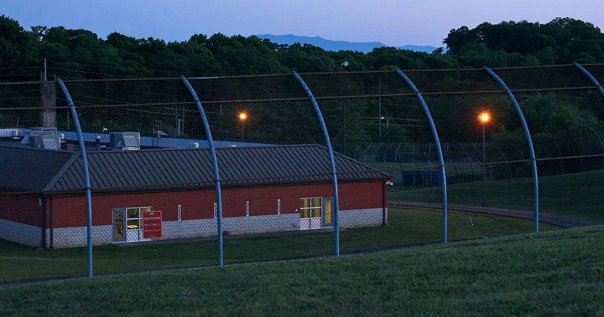 A fenced prison perimeter with curved blue posts, a red building, warning signs, and lights at dusk.
