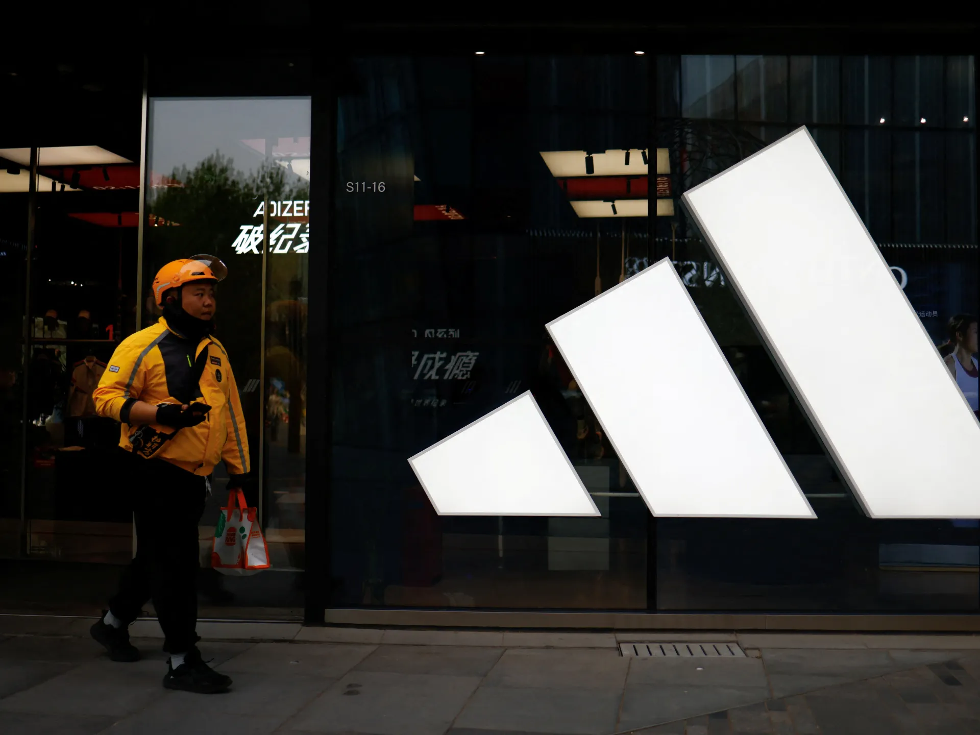 Delivery worker walks past a large illuminated Adidas logo outside a storefront at dusk.