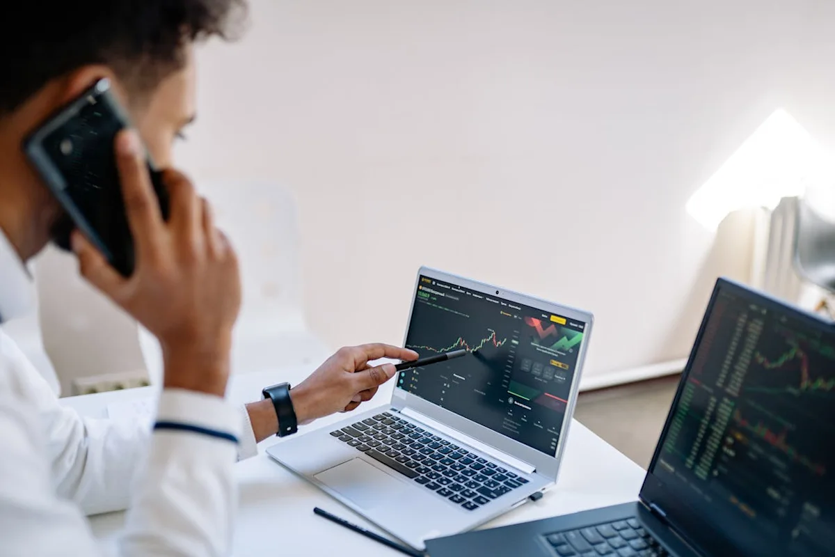 Two professionals examine financial charts on laptops while one speaks on a smartphone.