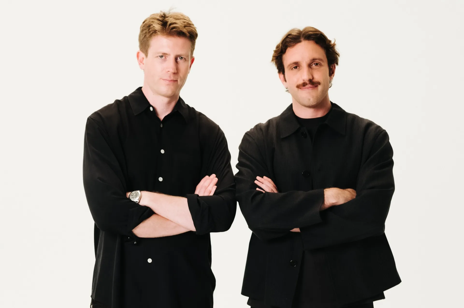 Two men in black outfits stand with arms crossed against a light background.