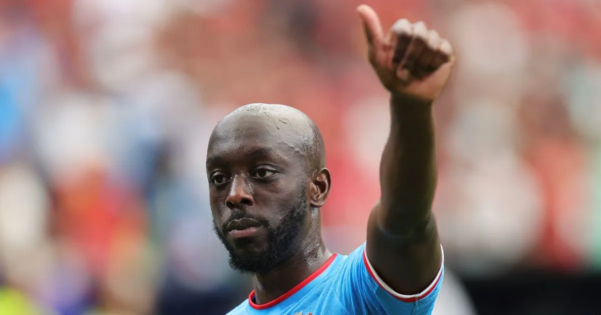 Bald, bearded footballer in a blue jersey raises his thumb against a blurred stadium crowd.