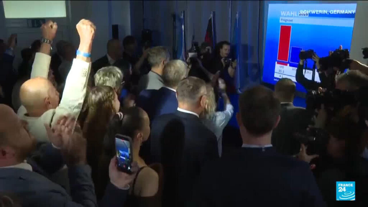 Crowd watches election results screen as photographers record people raising fists.