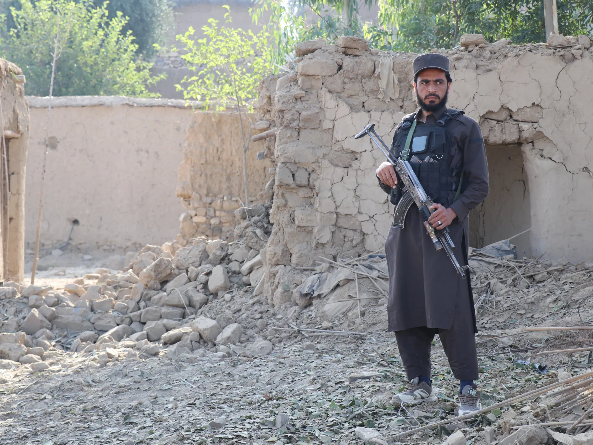 A man in dark clothing holds a rifle amid rubble beside a damaged earthen building.