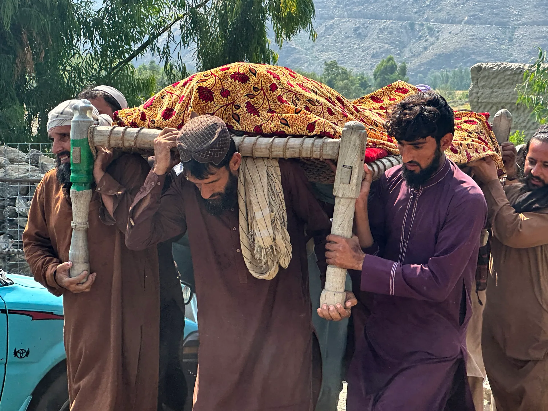 Men carry a cloth-covered stretcher outdoors against a mountainous backdrop.
