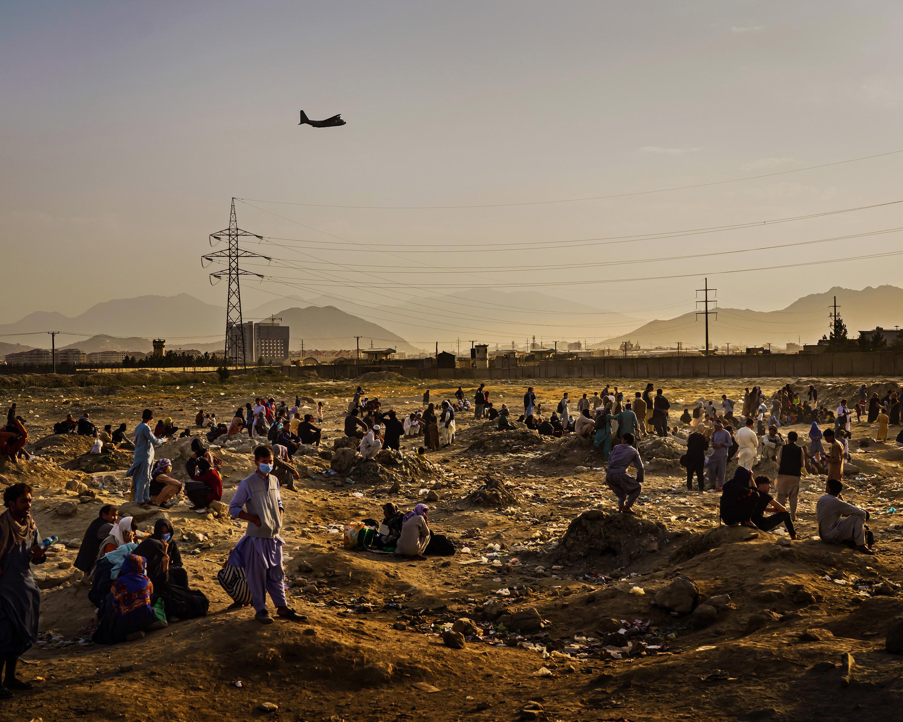Crowd gathers in a dusty field beneath power lines, mountains, and a passing aircraft.