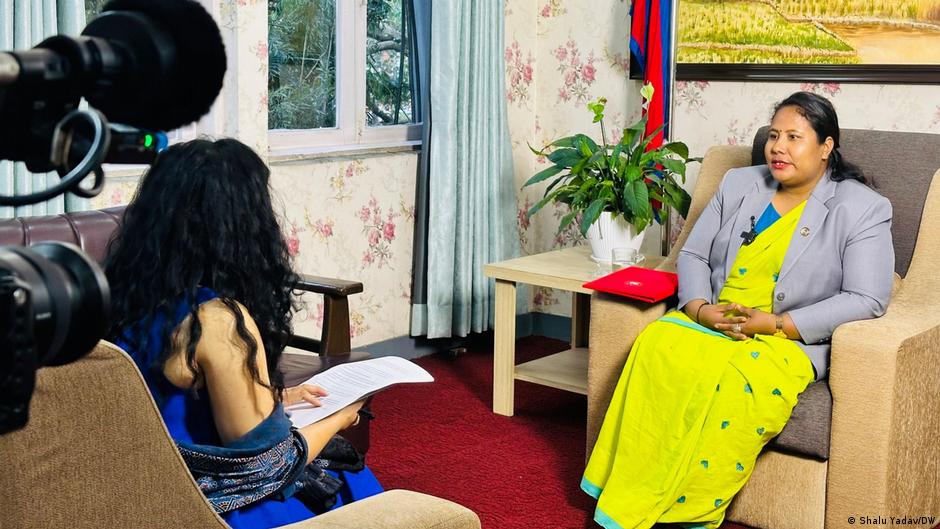Woman in yellow sari and blazer interviewed indoors as cameras record the conversation