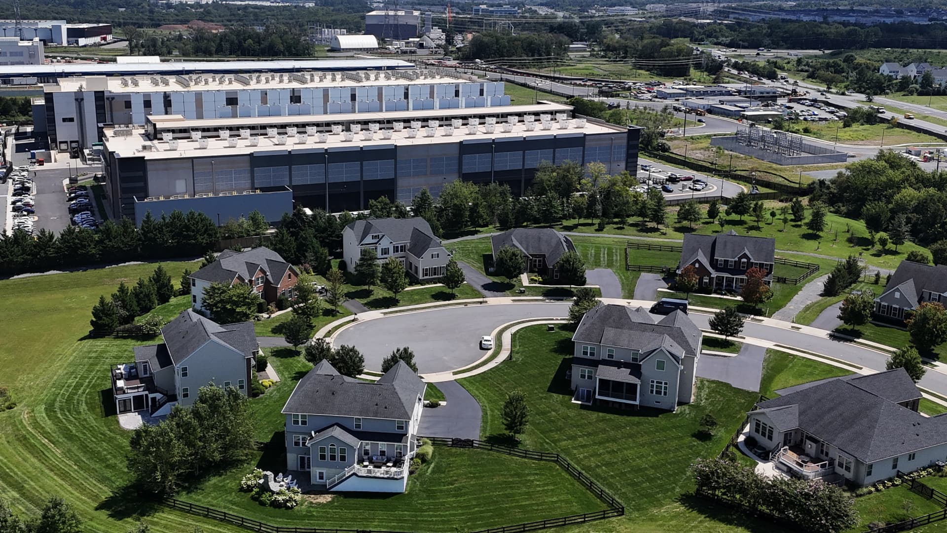 A large industrial data center stands behind landscaped suburban homes and green lawns.