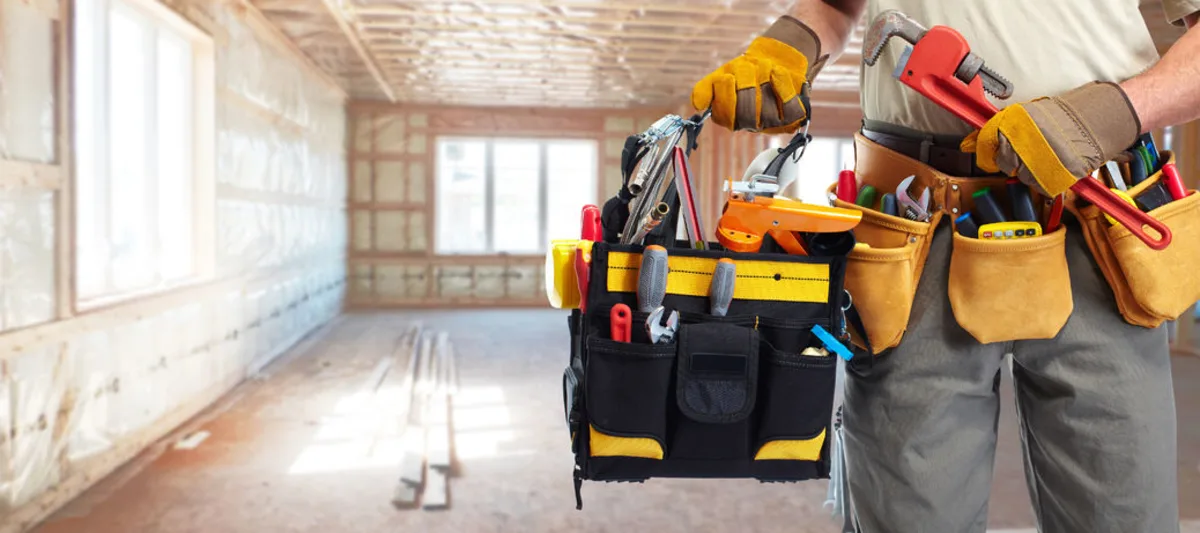 Construction worker holding a tool bag and wearing a utility belt inside an unfinished building.