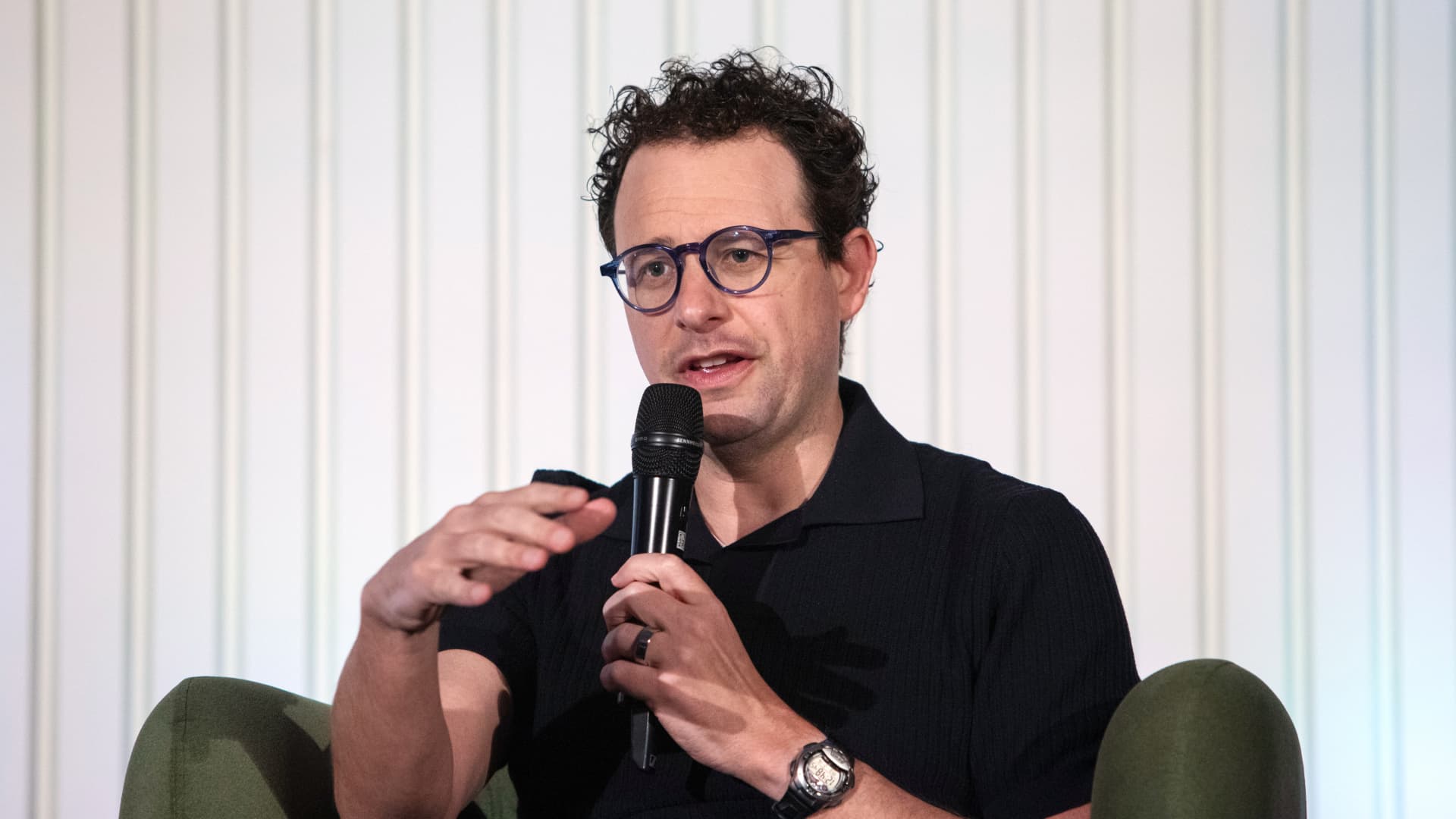 Curly-haired man wearing glasses speaks into a microphone while gesturing in a green armchair.