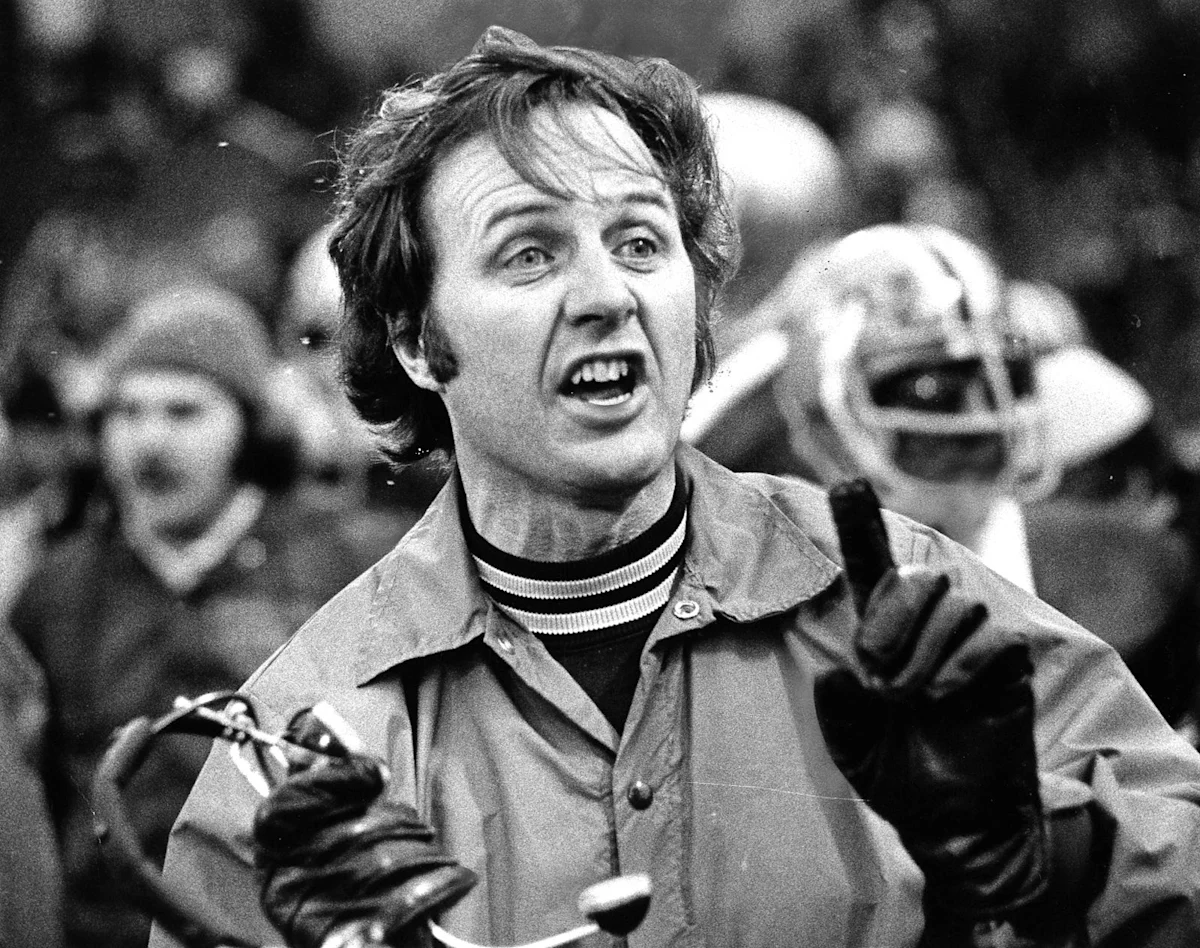 Black-and-white image of a shouting man in gloves gesturing amid a crowded football sideline