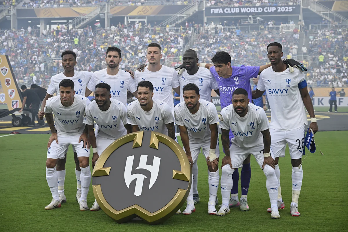 Al-Hilal football players pose together on the pitch with a large club emblem.