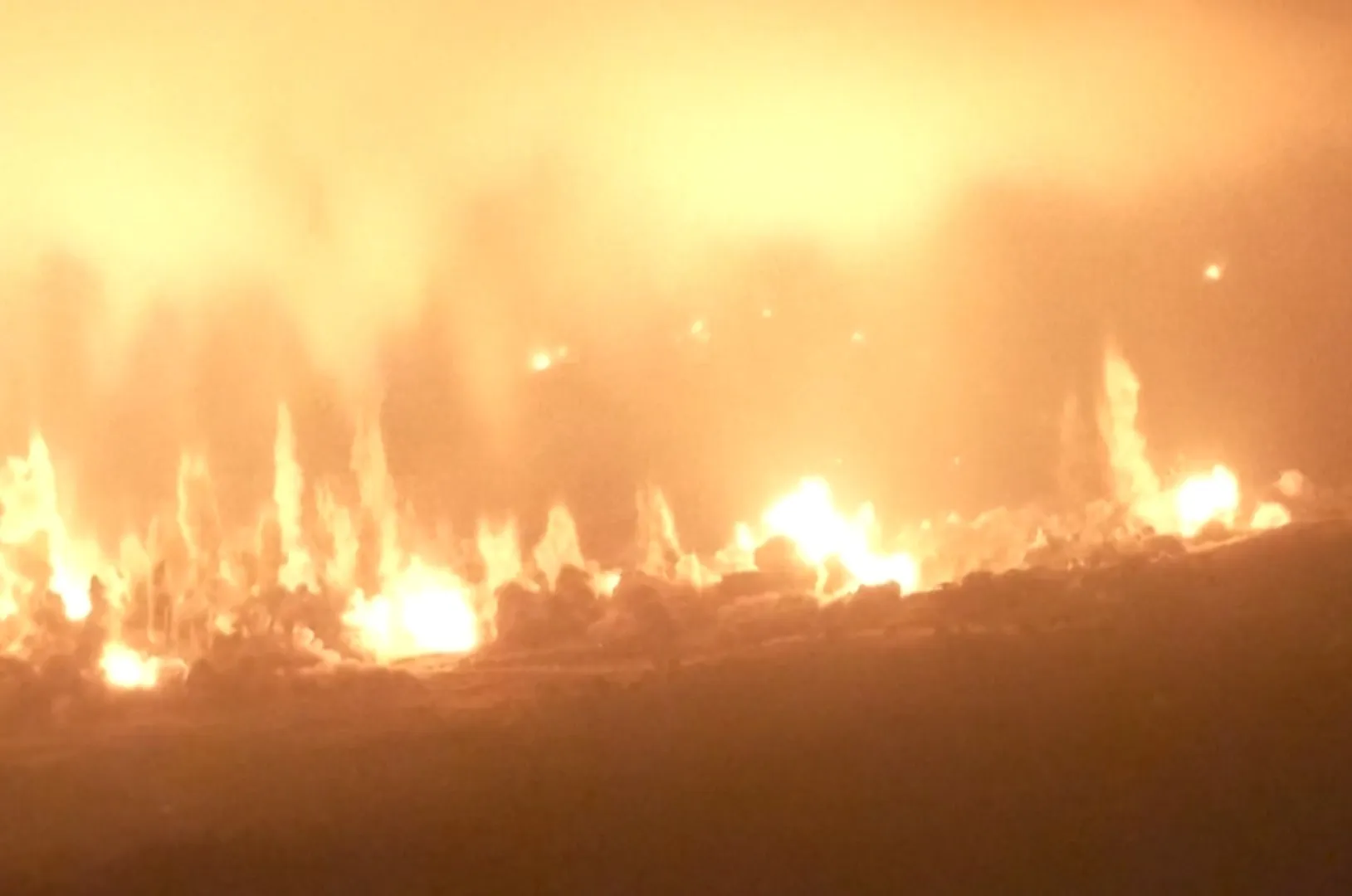 Multiple bright fires burn across a dark hillside at night, creating an intense orange glow.