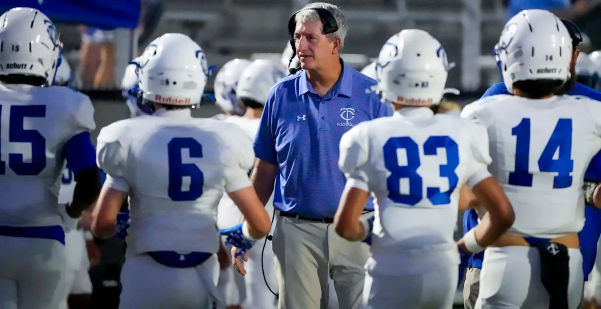 Football coach wearing headset stands among players in white and blue uniforms on sideline.