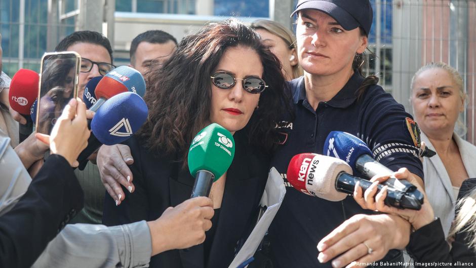 A woman in sunglasses is surrounded by reporters holding microphones outside a building.