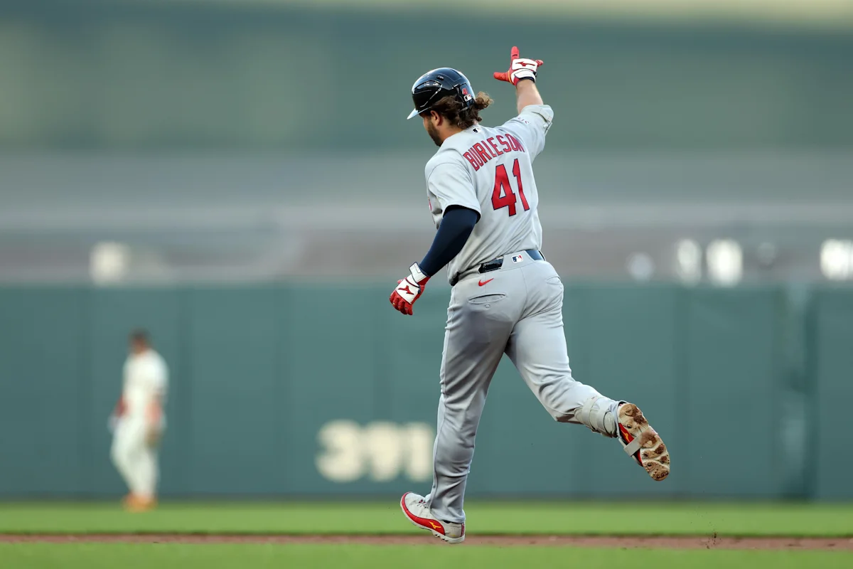Baseball player in Cardinals uniform runs with one arm raised after a hit.