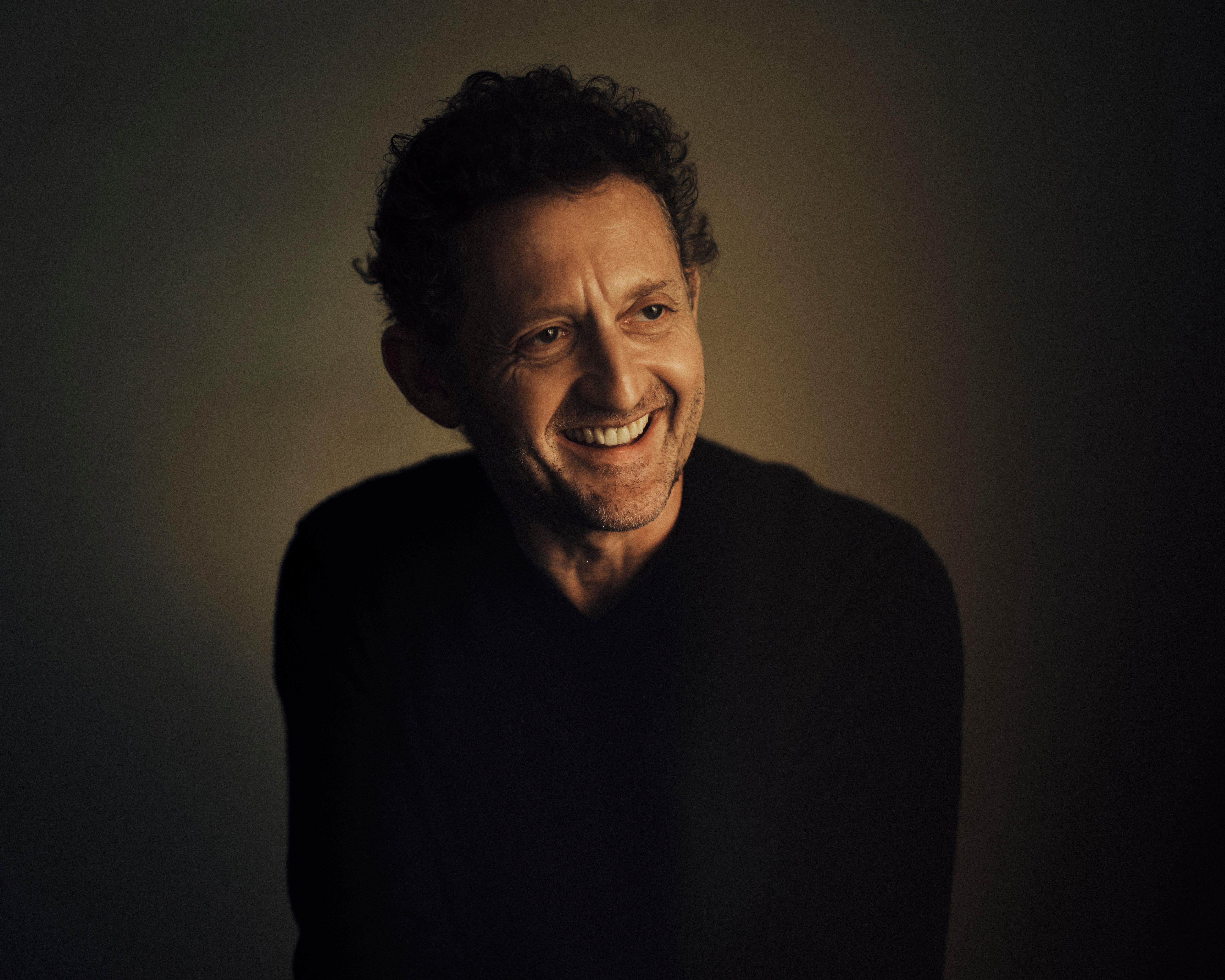 Smiling man with curly dark hair and stubble wearing a black sweater against a dimly lit wall