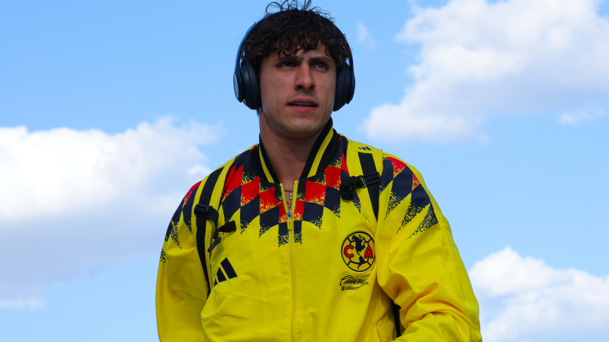 Man wearing Club América jacket, headphones, and backpack outdoors beneath a blue sky